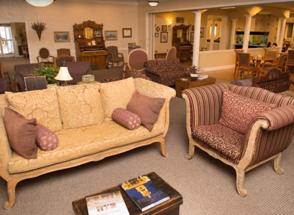 A cozy living room area in a senior living facility featuring a beige patterned sofa with purple and patterned cushions, a striped armchair with matching cushions, a small wooden coffee table with books, and additional seating and decor in the background including vintage furniture and a fish tank.
