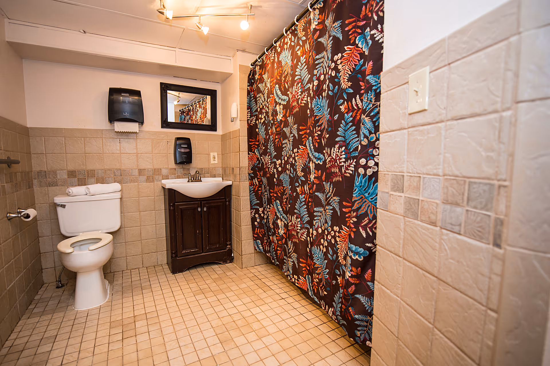 A clean bathroom with beige tiled walls and floor, featuring a white toilet with folded towels on top, a dark wooden vanity with a white sink, a mirror above the sink, a black paper towel dispenser, a soap dispenser, and a shower area with a colorful floral-patterned shower curtain.