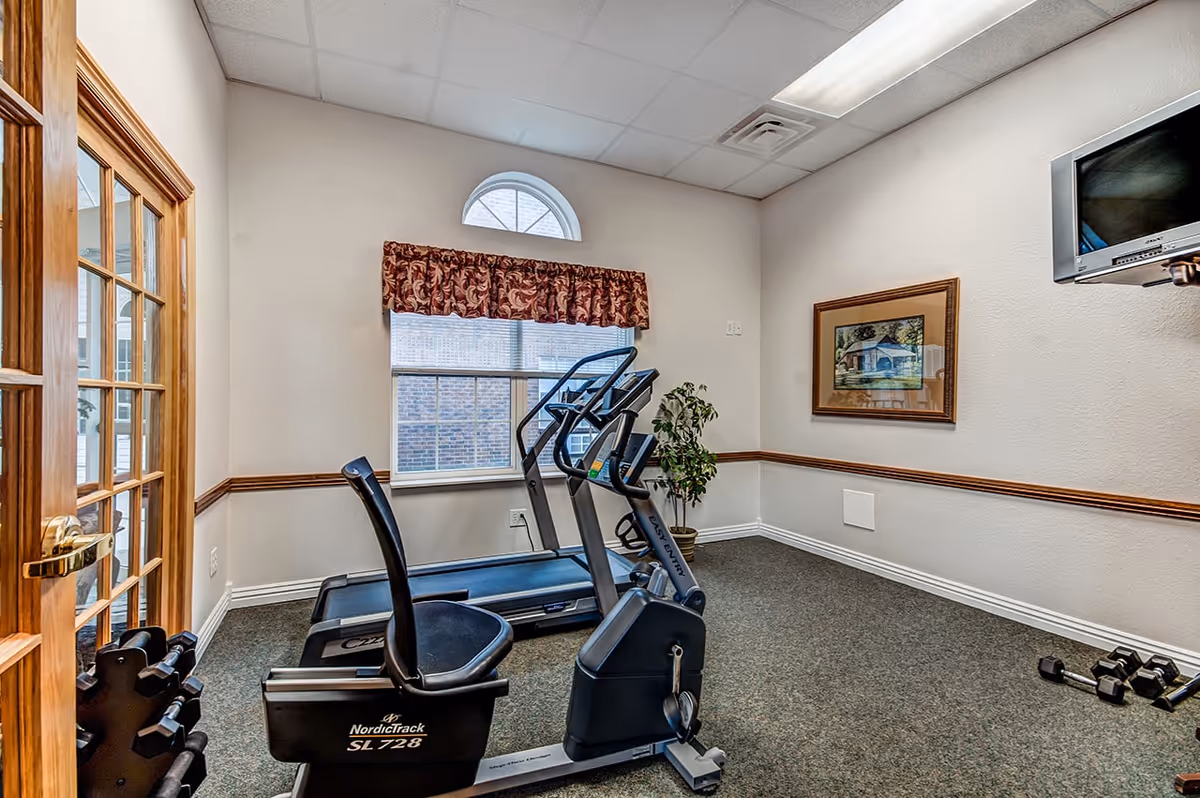 Small exercise room with a recumbent stationary bike, treadmill, dumbbells on the floor, a framed picture on the wall, a window with a half-circle top and floral valance, and a wall-mounted television.