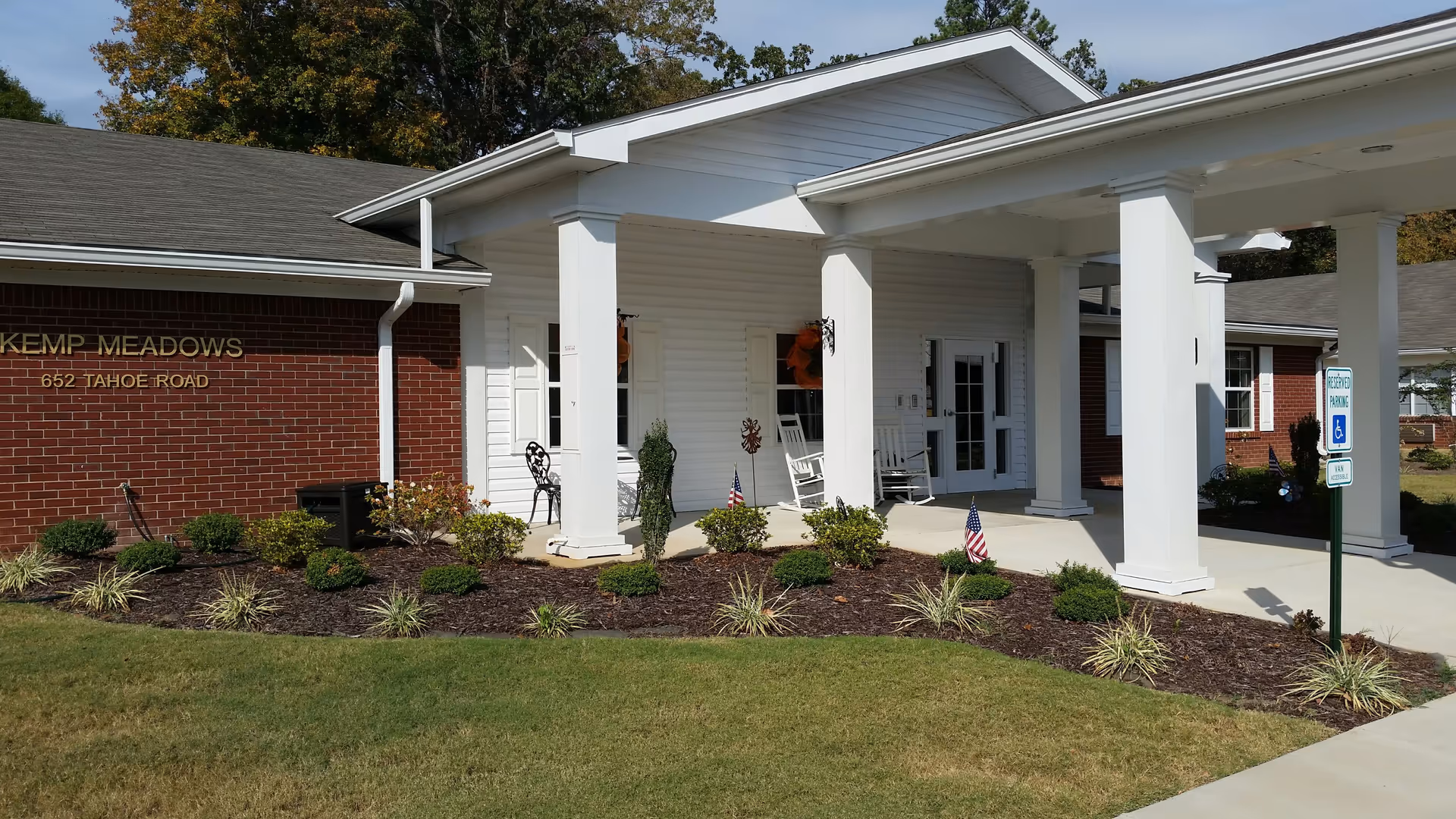 Front exterior view of Kemp Meadows Assisted Living facility showing a brick and white siding building with a covered entrance supported by white columns. There are rocking chairs on the porch, small bushes and plants in the landscaped area, and a reserved parking sign for handicapped access near the sidewalk.