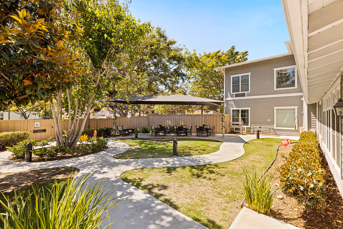 Sunny landscaped courtyard with winding concrete paths, shaded seating area under a canopy, and an adjacent two-story building.