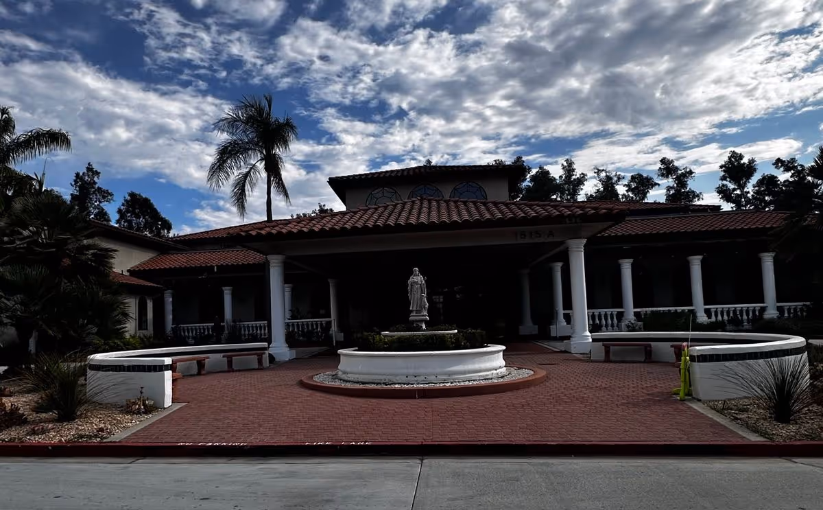 Front entrance of a Spanish-style building with a tiled roof, columns, a central circular fountain and statue, and palm trees under a partly cloudy sky.