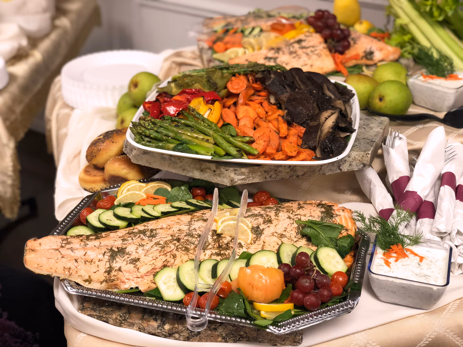A buffet table with a large platter of cooked salmon garnished with cucumber slices, grape tomatoes, grapes, lemon slices, and greens. Above it, a tray with grilled vegetables including asparagus, carrots, mushrooms, and bell peppers. There are also bagels, green apples, a bowl of creamy dip with herbs and shredded carrots, and neatly rolled napkins with purple napkin rings.
