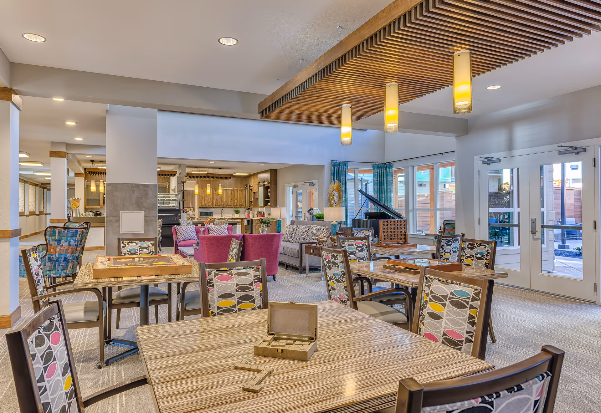 Bright and spacious common area in an assisted living facility featuring multiple tables and chairs with colorful patterned upholstery, a seating area with purple and gray sofas, a wooden ceiling accent with hanging pendant lights, large windows with blue curtains, and a grand piano near the glass doors leading outside.