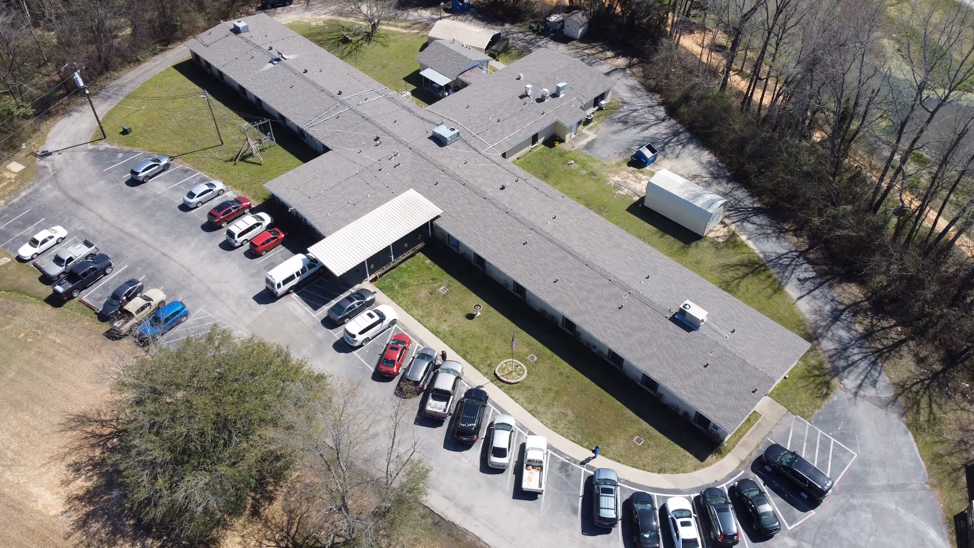 Aerial view of Lakeview Rehabilitation and Healthcare Center showing a single-story building with a cross-shaped layout surrounded by parking spaces with multiple cars parked. The building is situated in a green area with trees and open grassy spaces around it.