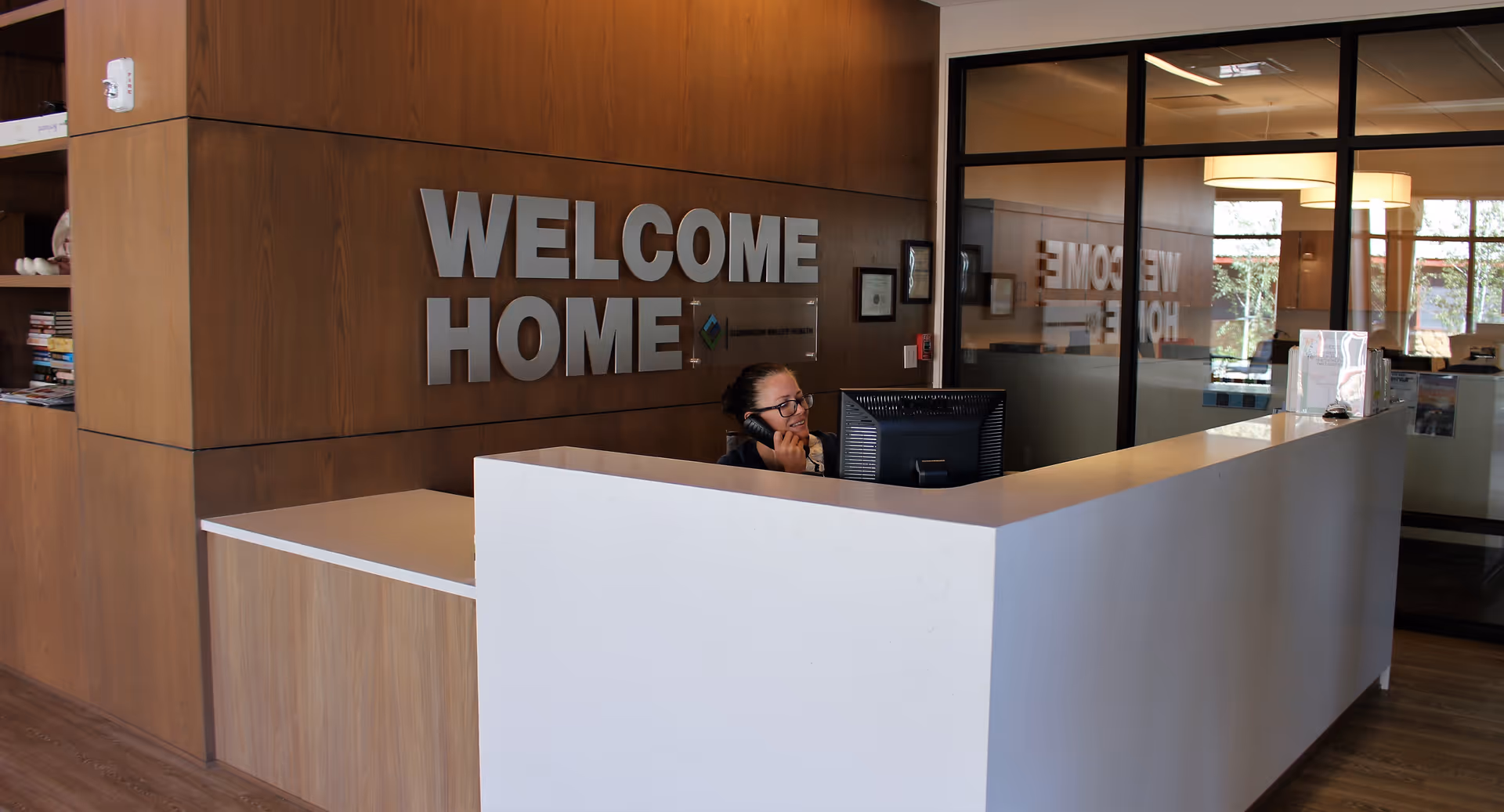 Reception area with a 'WELCOME HOME' sign on a wood wall and a staff member speaking on the phone behind a white front desk.