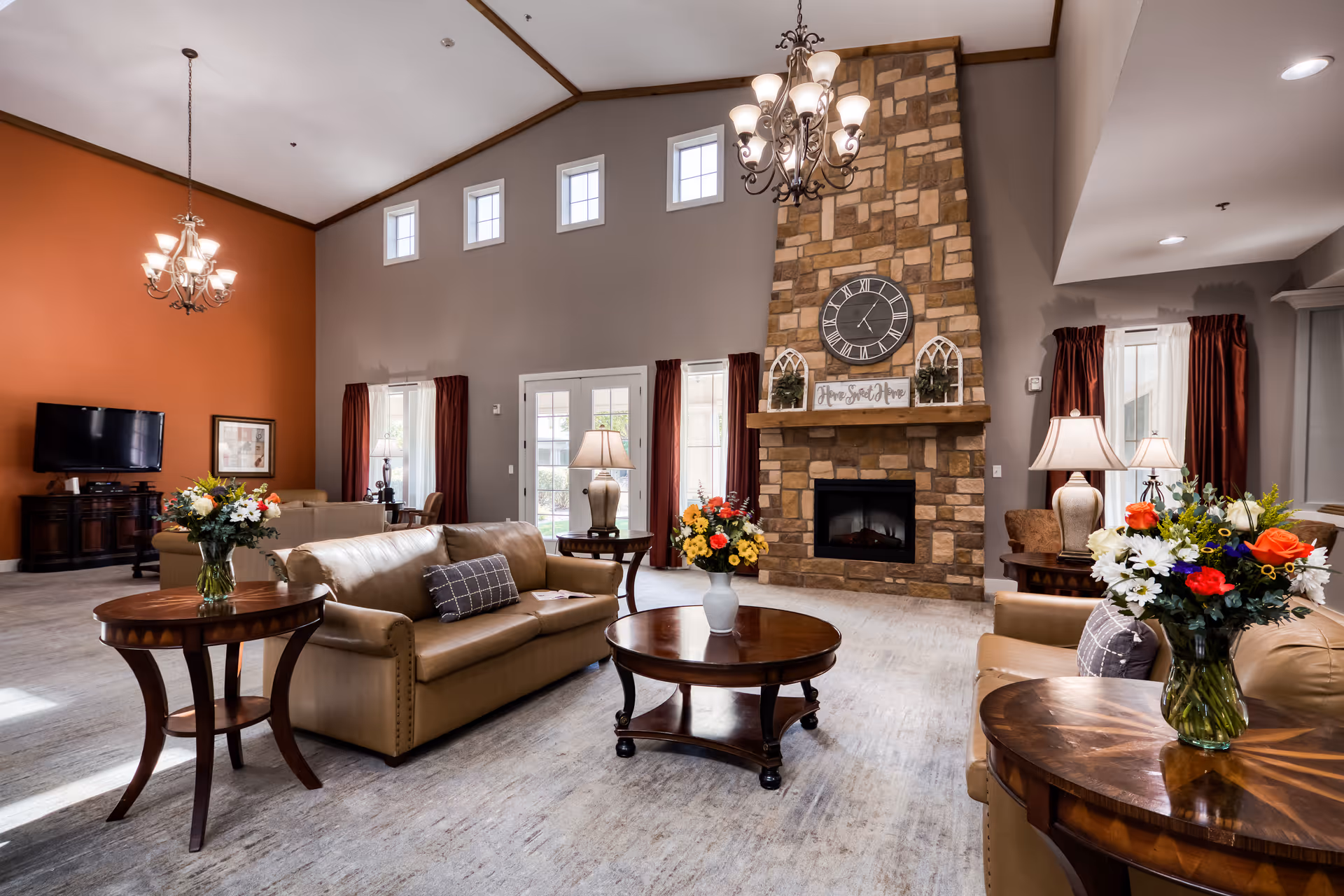 A spacious and well-lit living room in an assisted living facility featuring beige leather sofas, wooden coffee and side tables with floral arrangements, a stone fireplace with a large clock and a 'Home Sweet Home' sign above it, multiple windows with red curtains, and two chandeliers hanging from a high ceiling.