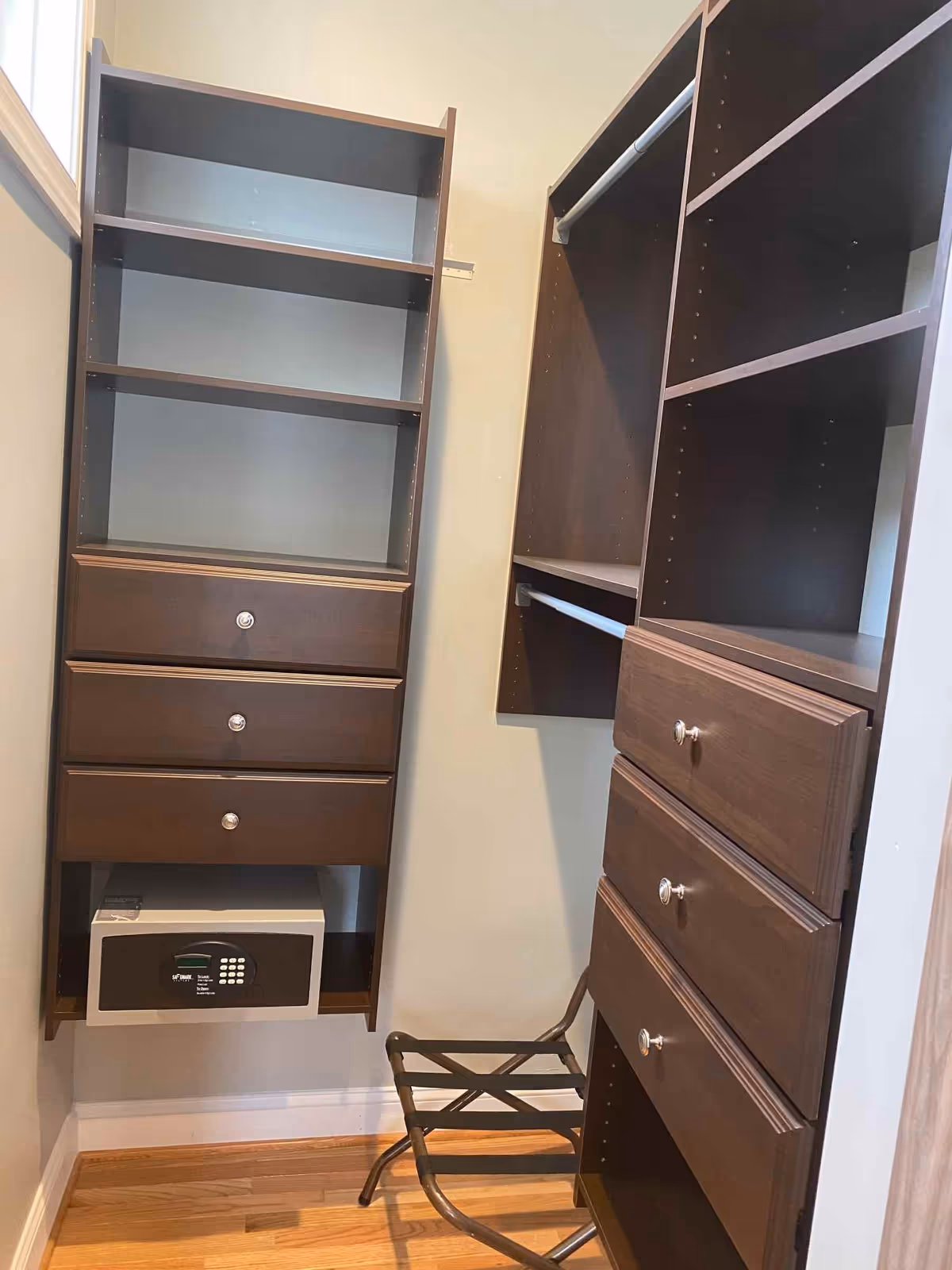 Interior view of a walk-in closet with dark wood shelving units, drawers, hanging rods, a small safe, and a luggage rack on a wooden floor.