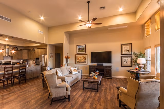 A spacious and well-lit living room area with beige walls and wooden flooring. The room features a beige sofa, two armchairs, a wooden coffee table, and a flat-screen TV mounted on a dark wooden TV stand. There are framed artworks on the wall behind the TV and a ceiling fan with lights. To the left, an open kitchen area with wooden cabinets, stainless steel appliances, and a breakfast bar with chairs is visible. Large windows with blinds allow natural light to fill the room.