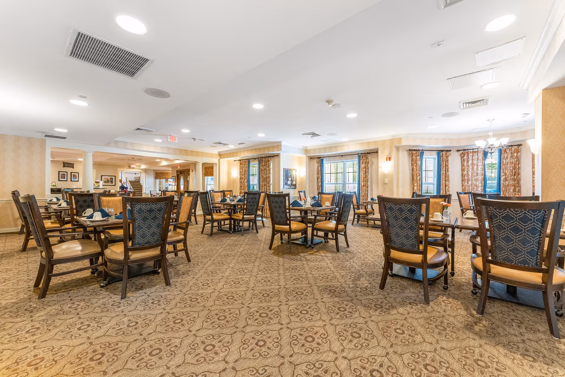 A spacious dining room with multiple wooden tables and chairs arranged neatly. The room has patterned carpet flooring, large windows with floral curtains, and soft lighting from ceiling fixtures. The walls are decorated with framed pictures and the overall ambiance is warm and inviting.