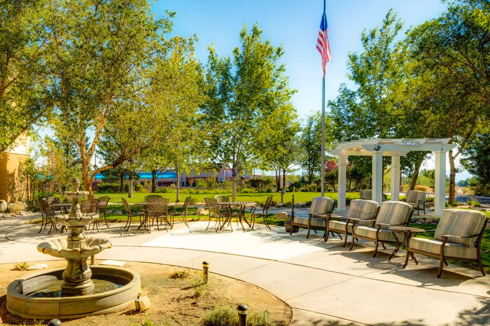 Outdoor seating area at The Seasons of Reno featuring a stone fountain in the foreground, several metal tables and chairs, cushioned armchairs arranged in a row, a white pergola, green trees, and an American flag on a flagpole under a clear blue sky.