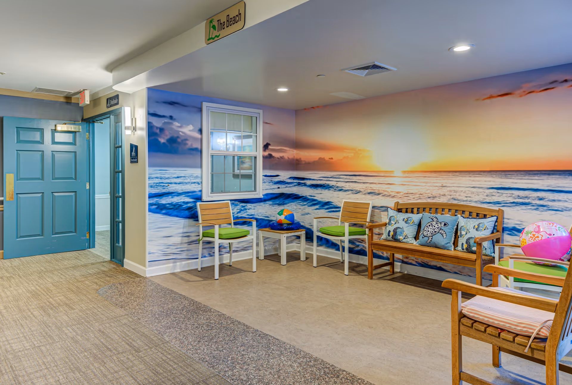 A seating area in a senior living facility with a beach-themed mural showing an ocean sunset on the wall. The area includes wooden and metal chairs with cushions, a small table with a colorful beach ball, and decorative pillows with sea turtle designs on a wooden bench. There is a blue door and hallway visible to the left, and a sign above the seating area reads 'The Beach'.