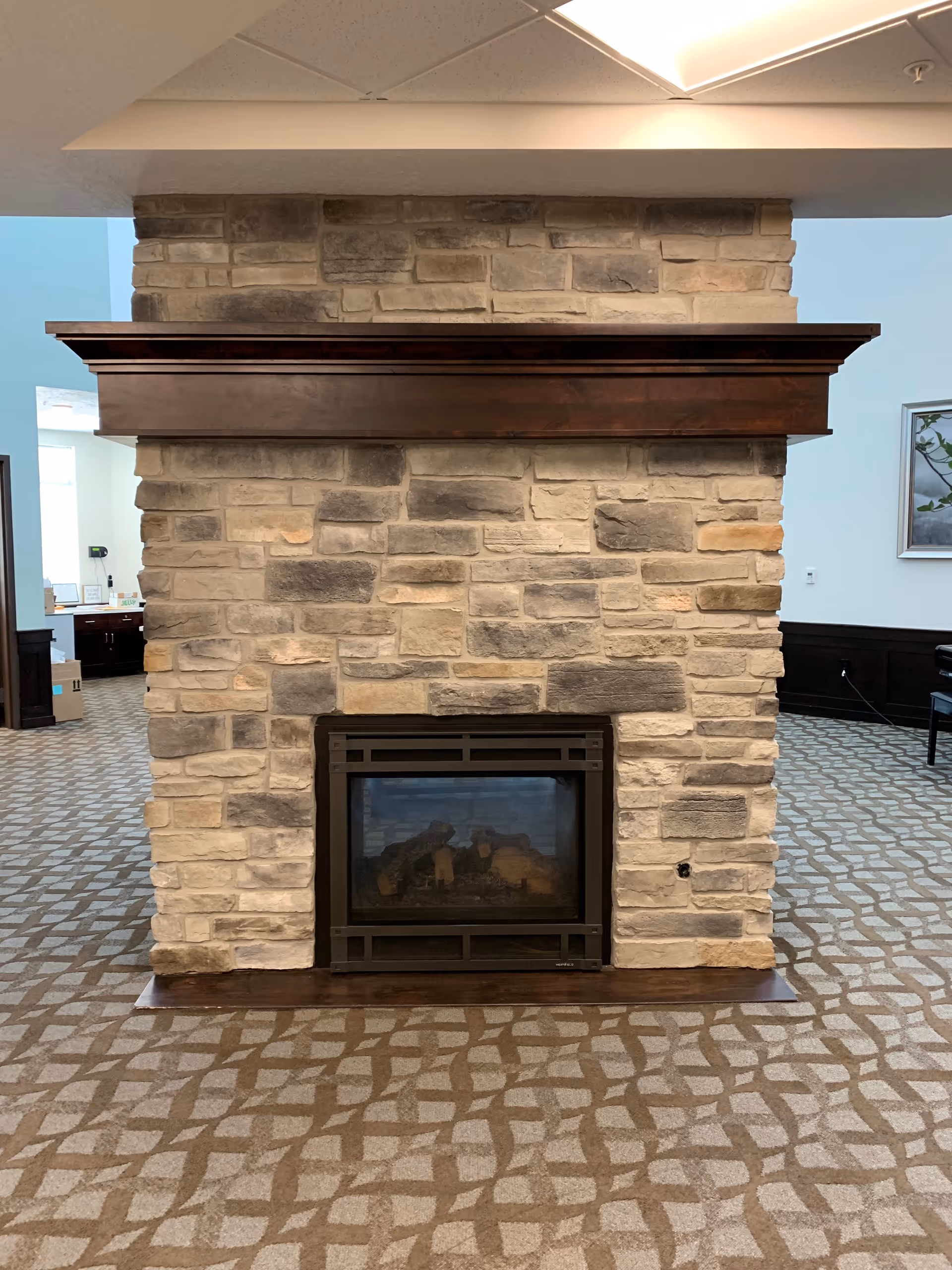 A stone fireplace with a dark wooden mantel in a room with patterned carpet. The fireplace is built with light and dark beige stones and has a glass front. The room has light blue walls and some furniture is partially visible in the background.
