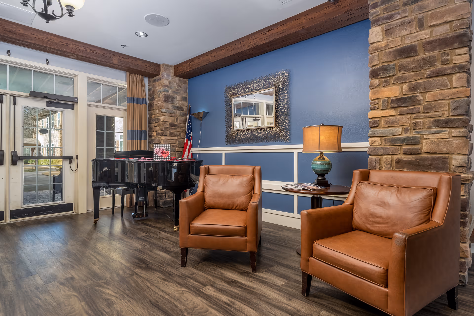 A cozy sitting area with two brown leather armchairs and a small round table with a lamp and magazines. Behind the chairs is a blue wall with white trim and a decorative mirror. To the left, there is a black grand piano near a set of glass double doors with windows, and an American flag is placed in the corner. The floor is wood with a natural finish, and the room features exposed stone columns and wooden beams on the ceiling.