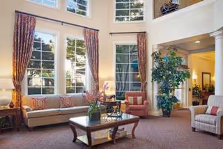 A spacious living room with high ceilings and large windows letting in natural light. The room features a beige sofa with patterned cushions, two armchairs, a wooden coffee table with a flower arrangement, a tall indoor plant, and decorative curtains on the windows.