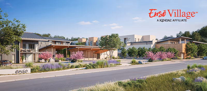 Exterior view of Ensō Village Preview Center showing modern buildings surrounded by landscaped greenery, trees, and flowering plants along a paved road under a clear sky.