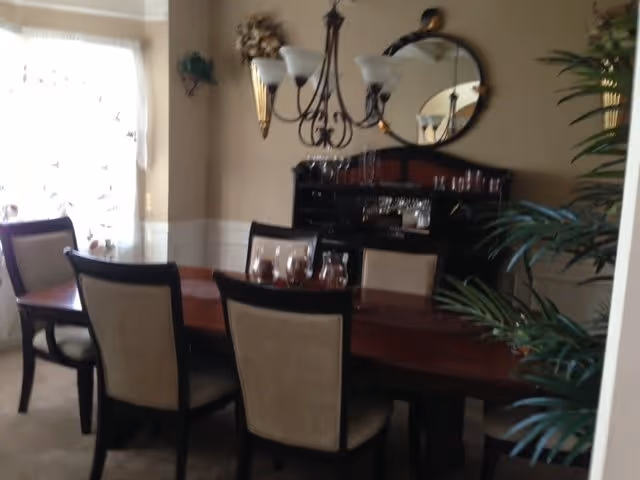 A dining room with a wooden dining table surrounded by six chairs with beige upholstery. A chandelier with five lights hangs above the table. There is a black sideboard with glassware and a round mirror on the wall above it. A large window with sheer curtains lets in natural light, and there are plants in the room adding greenery.