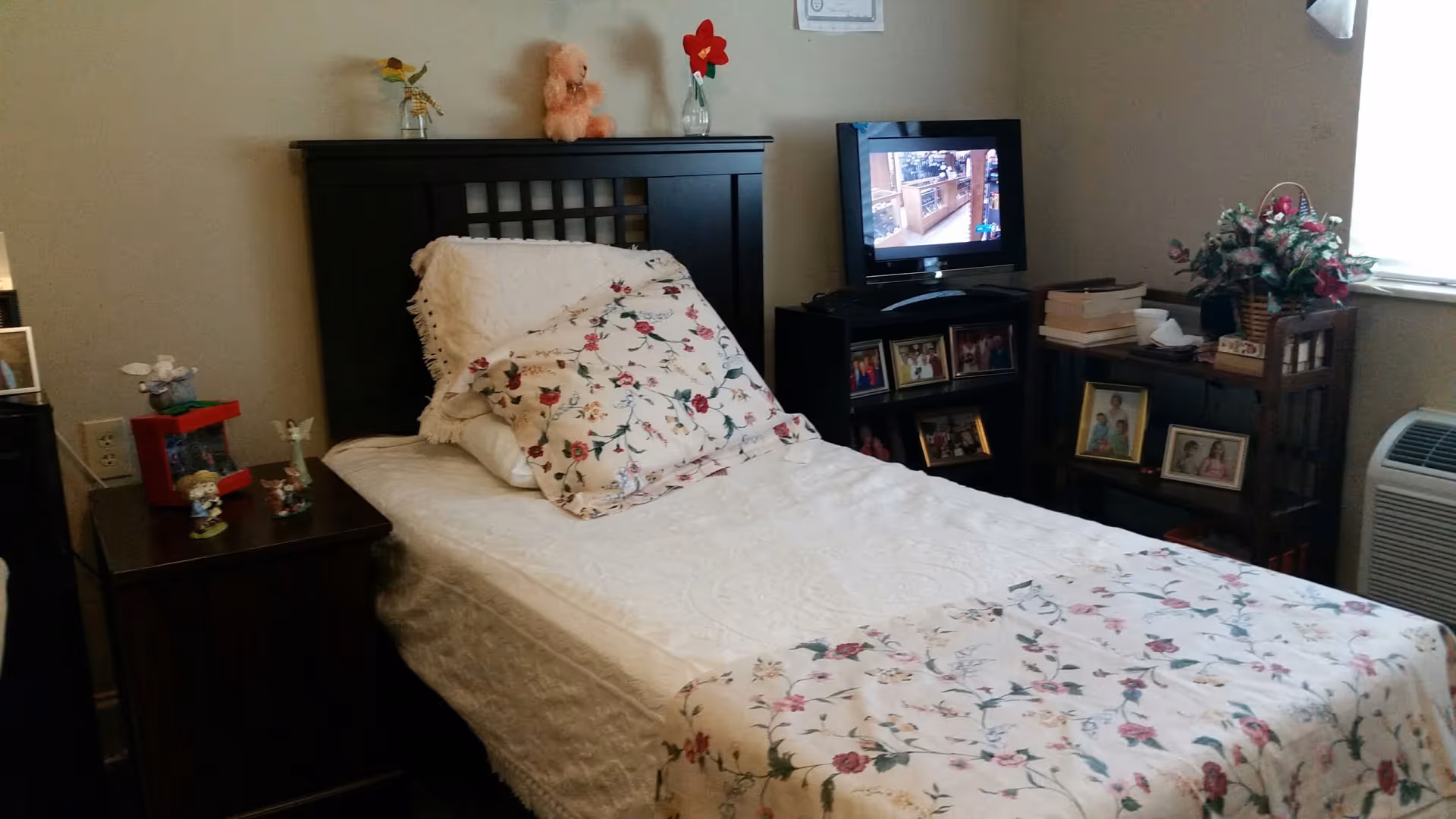 A cozy bedroom with a single bed featuring floral patterned bedding and pillows. The bed has a dark wooden headboard. Next to the bed is a small dark wooden nightstand with decorative items. Across from the bed is a small TV on a stand, with framed photos and books on shelves below and beside it. A window with blinds and a flower arrangement on a small table are also visible.