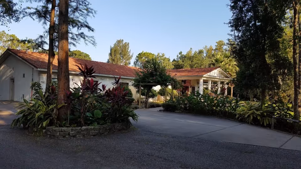 Exterior view of East Lake Manor Assisted Living Facility showing a single-story building with a red-tiled roof surrounded by trees and landscaped plants. There is a driveway leading to the entrance, which has a covered porch with white columns and an American flag hanging.