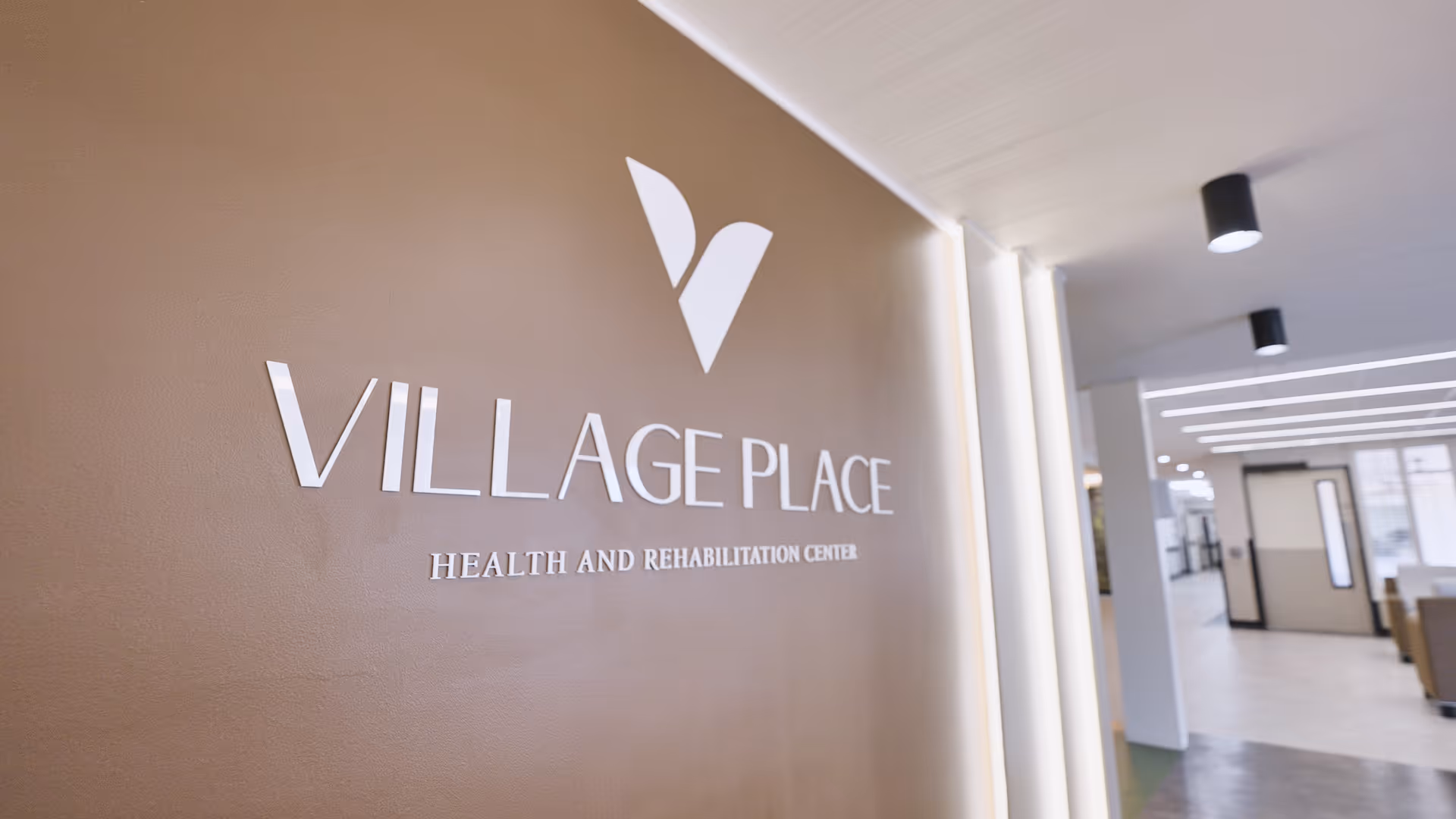 Interior view of a hallway in Village Place Health and Rehabilitation Center with the facility's name and logo displayed on a brown wall. The hallway is well-lit with modern ceiling lights and has a clean, spacious design.
