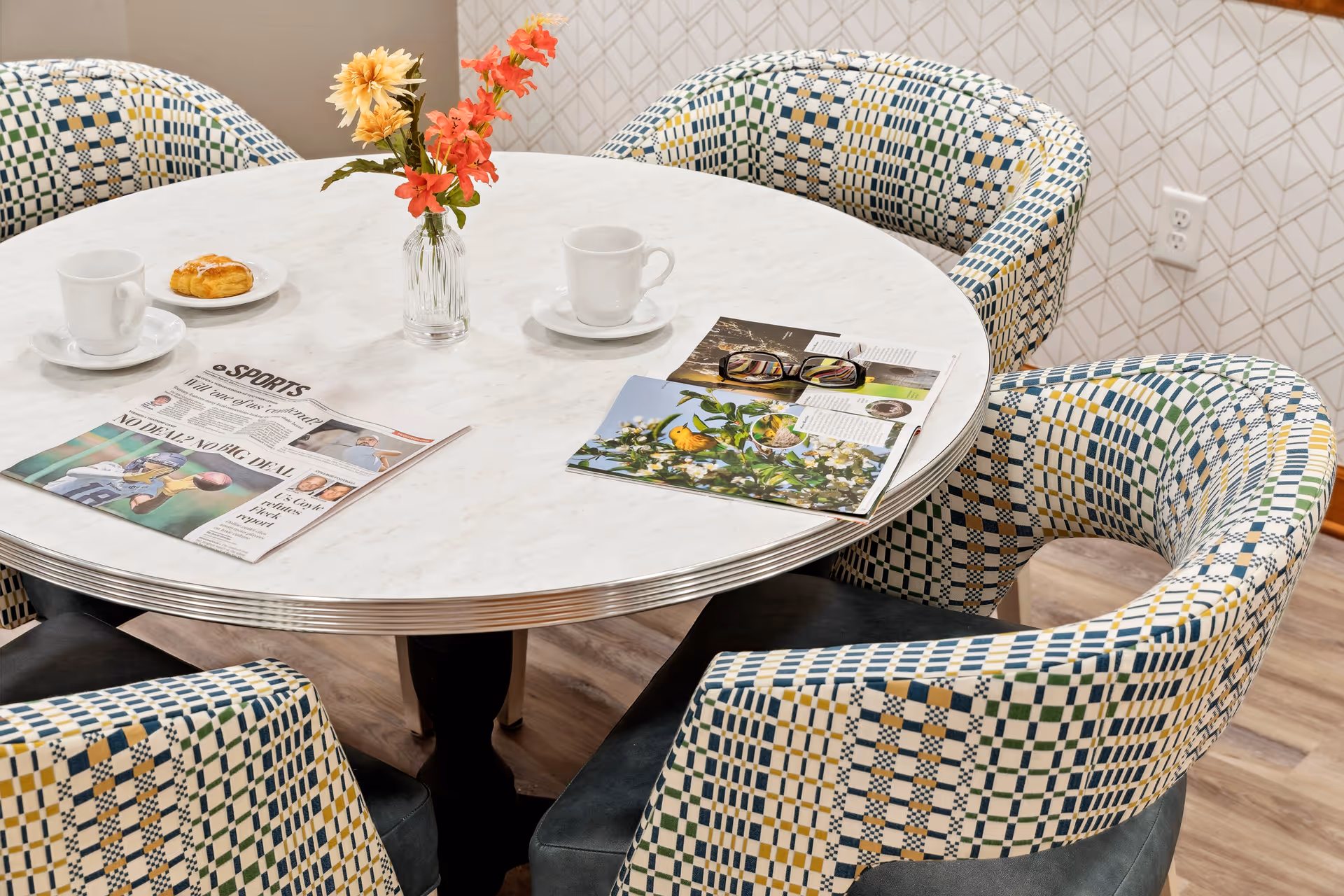 A round white marble table with a small vase of orange and yellow flowers in the center, surrounded by four patterned upholstered chairs. On the table are two white coffee cups with saucers, a pastry on a plate, a newspaper, and an open magazine with reading glasses resting on it. The background shows a wall with a geometric pattern and a power outlet.
