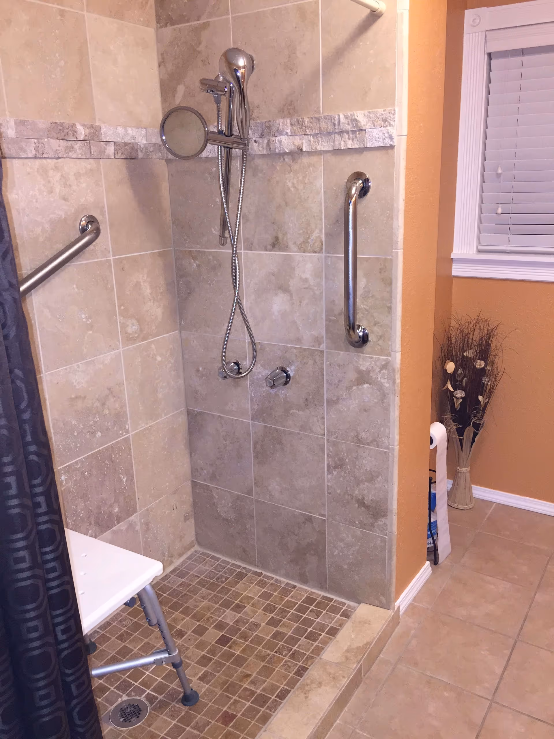 A tiled walk-in shower with a handheld showerhead, grab bars on the walls, and a white shower chair. The shower area has beige and brown tiles, and there is a small window with white blinds on the adjacent wall. A decorative vase with dried plants is placed near the window.