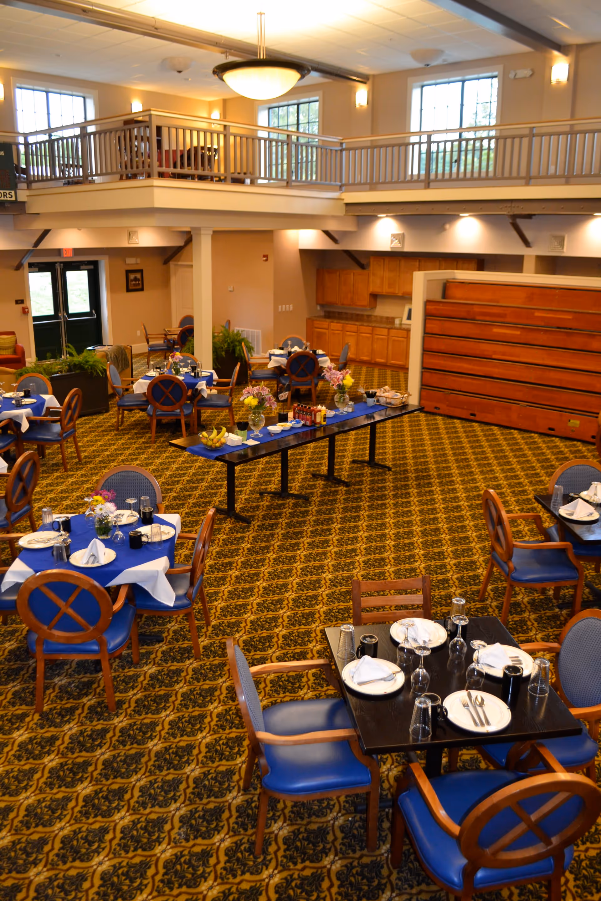 A spacious dining room in a senior living facility with multiple tables set with plates, glasses, and napkins. The room features blue cushioned chairs, a patterned carpet, and a long table in the center with flowers and condiments. There is a mezzanine level with railings and large windows letting in natural light.