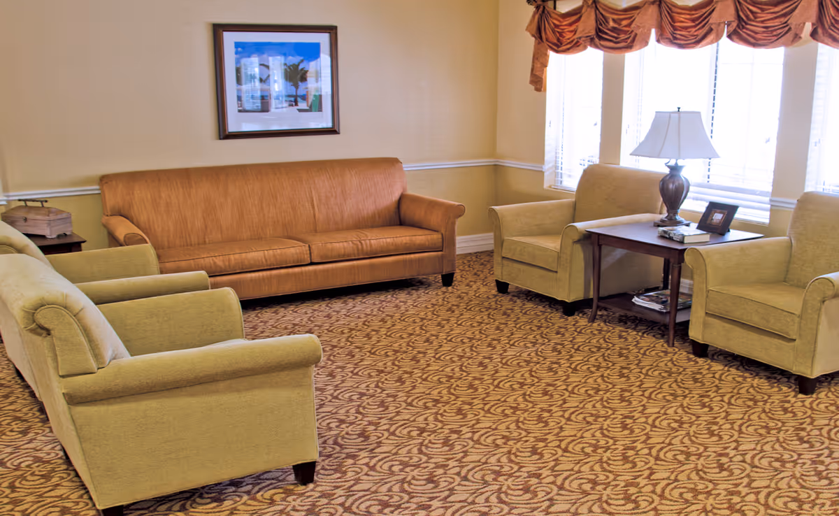 A cozy living room with a brown sofa, two beige armchairs, a wooden side table with a lamp, books, and a framed photo. The room has patterned carpet, beige walls with white trim, and a window with blinds and a brown valance.