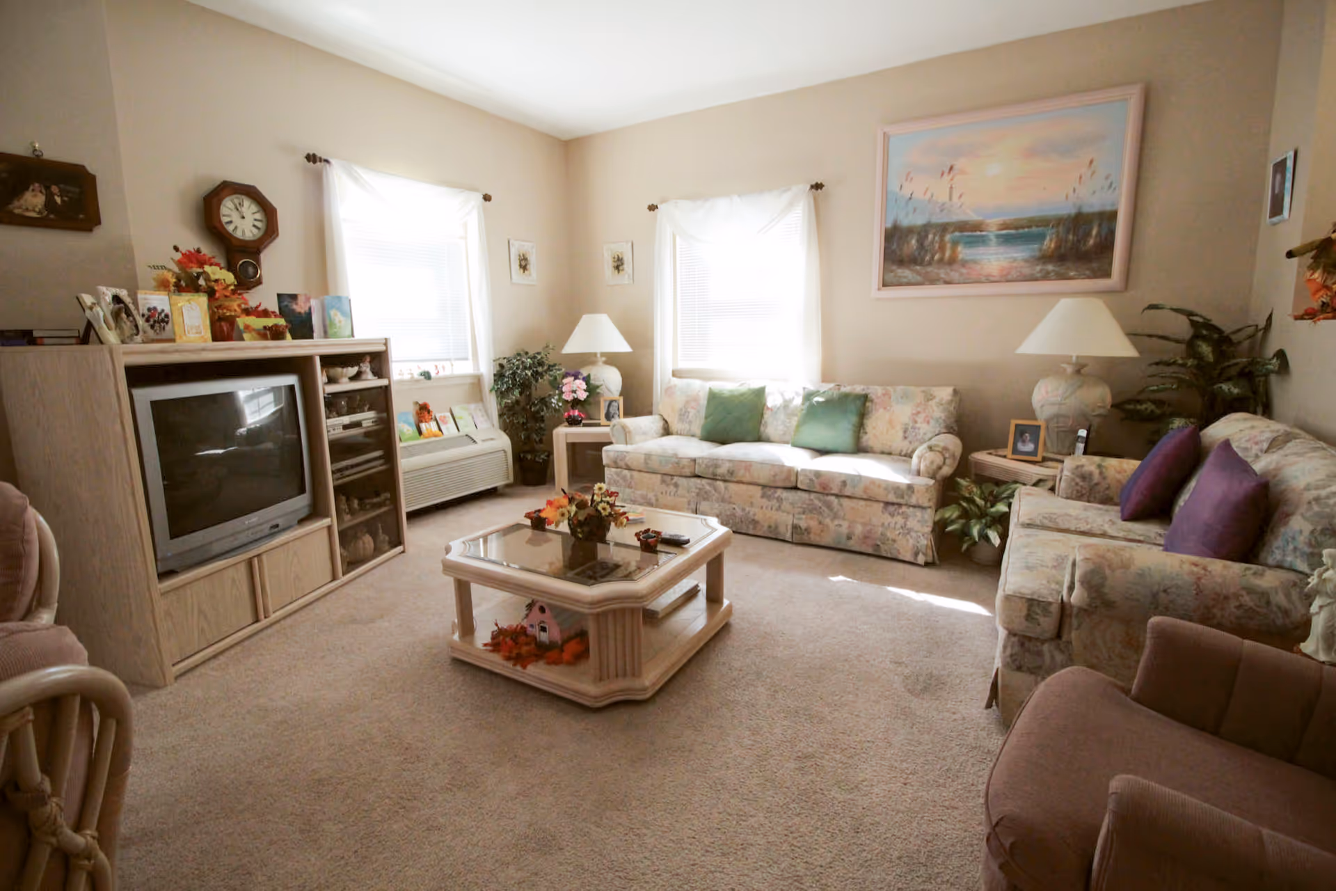 A cozy living room with two floral-patterned sofas adorned with green and purple pillows, a glass-top coffee table with decorative items, a wooden entertainment center with a TV and various decorations, two windows with white curtains, two table lamps on side tables, plants, and a large framed painting of a sunset over water on the wall.