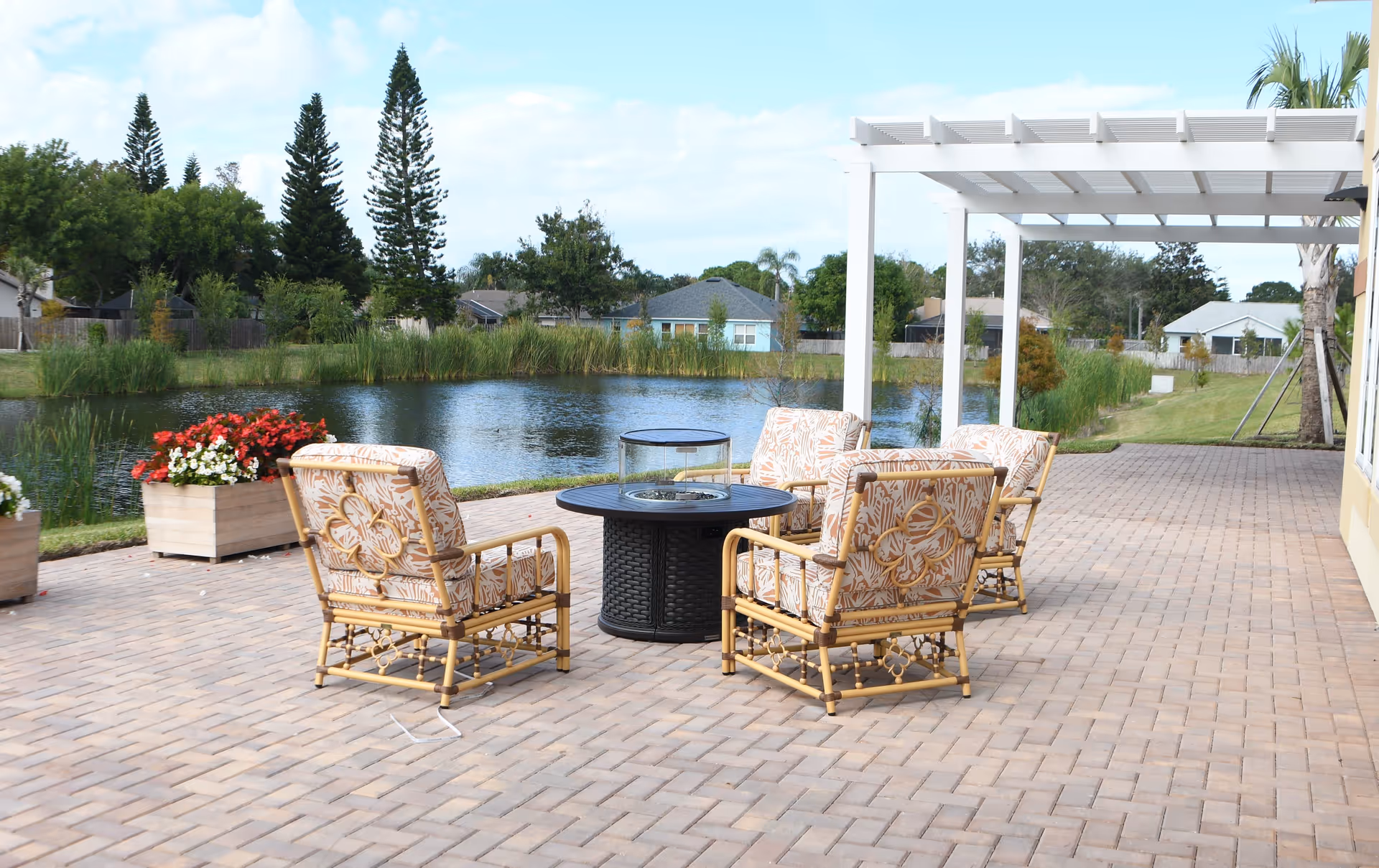 Outdoor patio area with four cushioned chairs arranged around a circular fire pit table, overlooking a pond with greenery and houses in the background under a partly cloudy sky.