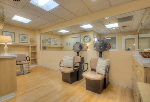 Interior of a salon area in a senior living facility with two beige salon chairs under hair dryers, a beige chair near a counter with a lamp, light wood flooring, and large mirrors on the walls reflecting the room.