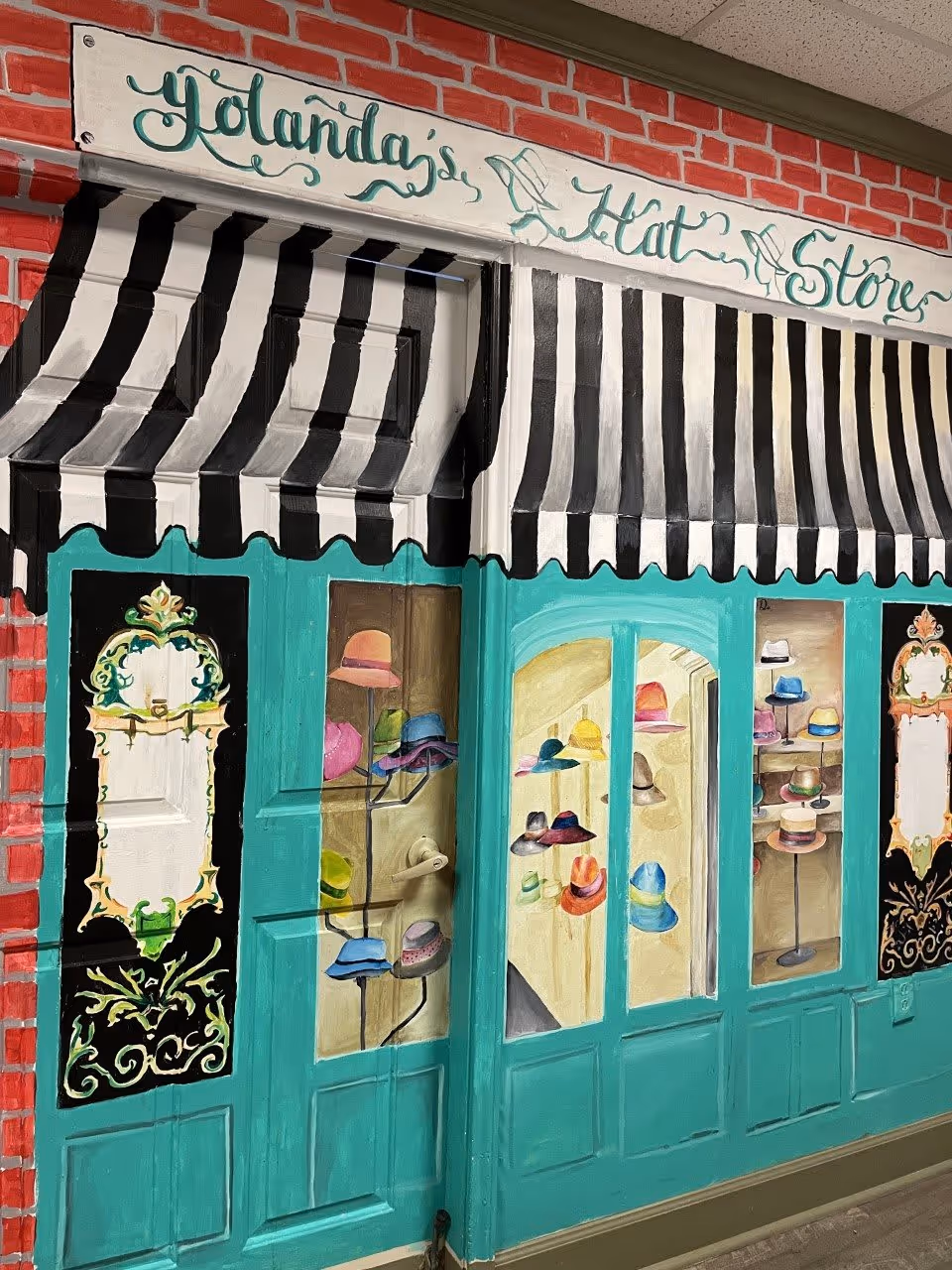 A painted mural on an interior wall depicting a storefront named 'Yolanda's Hat Store' with a black and white striped awning and colorful hats displayed in the windows. The mural includes decorative elements and a red brick pattern on the upper part of the wall.