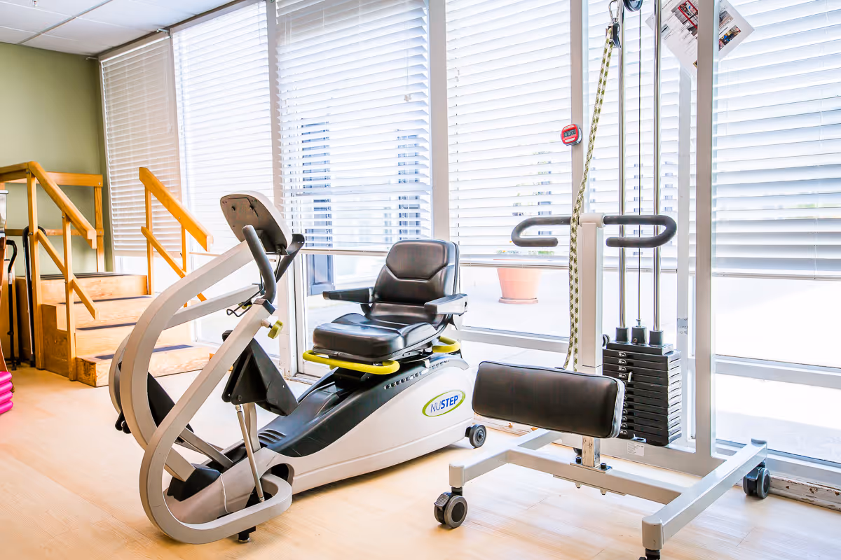 Exercise/rehab room with a recumbent NuStep bike and a cable weight machine by large windows with blinds.