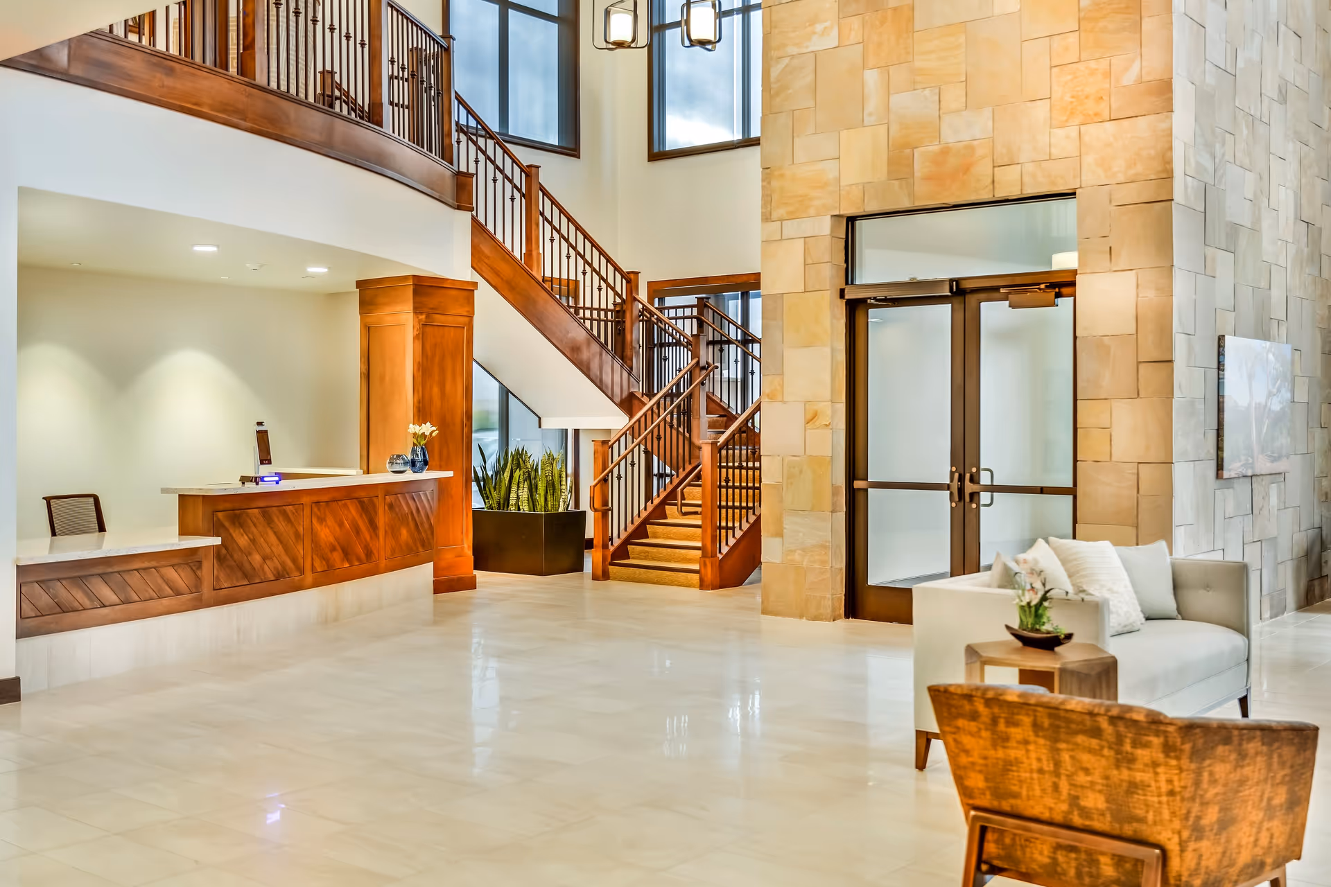 Bright and spacious lobby area with a wooden reception desk on the left, a staircase with wooden railings leading to an upper floor, large windows letting in natural light, a stone wall with double glass doors, and a seating area with a white couch and a wooden chair.