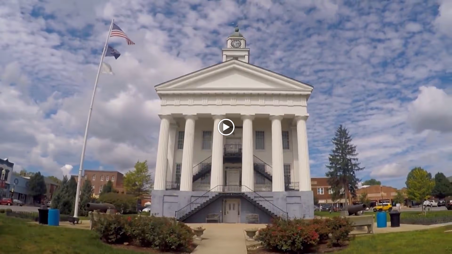 A large white classical building with tall columns and a clock tower on top, set against a partly cloudy sky. There are flags on a flagpole to the left and greenery including bushes and trees around the building. Nearby buildings and a street are visible in the background.