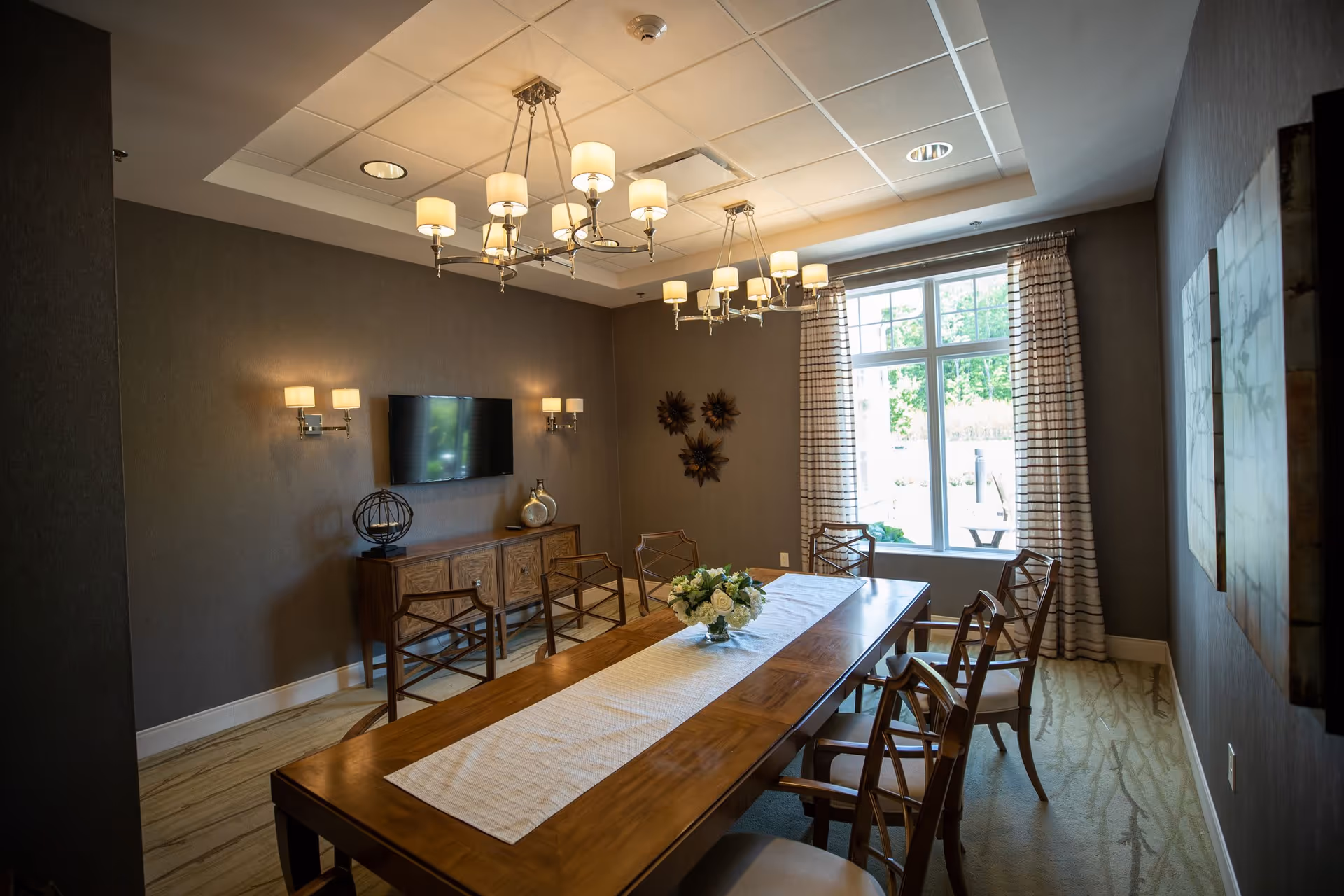 A well-lit dining room with a long wooden table covered by a white runner and a floral centerpiece. The room features eight wooden chairs, a large window with striped curtains allowing natural light, a wall-mounted flat-screen TV, two wall sconces, a wooden sideboard with decorative items, and two modern chandeliers hanging from the ceiling.