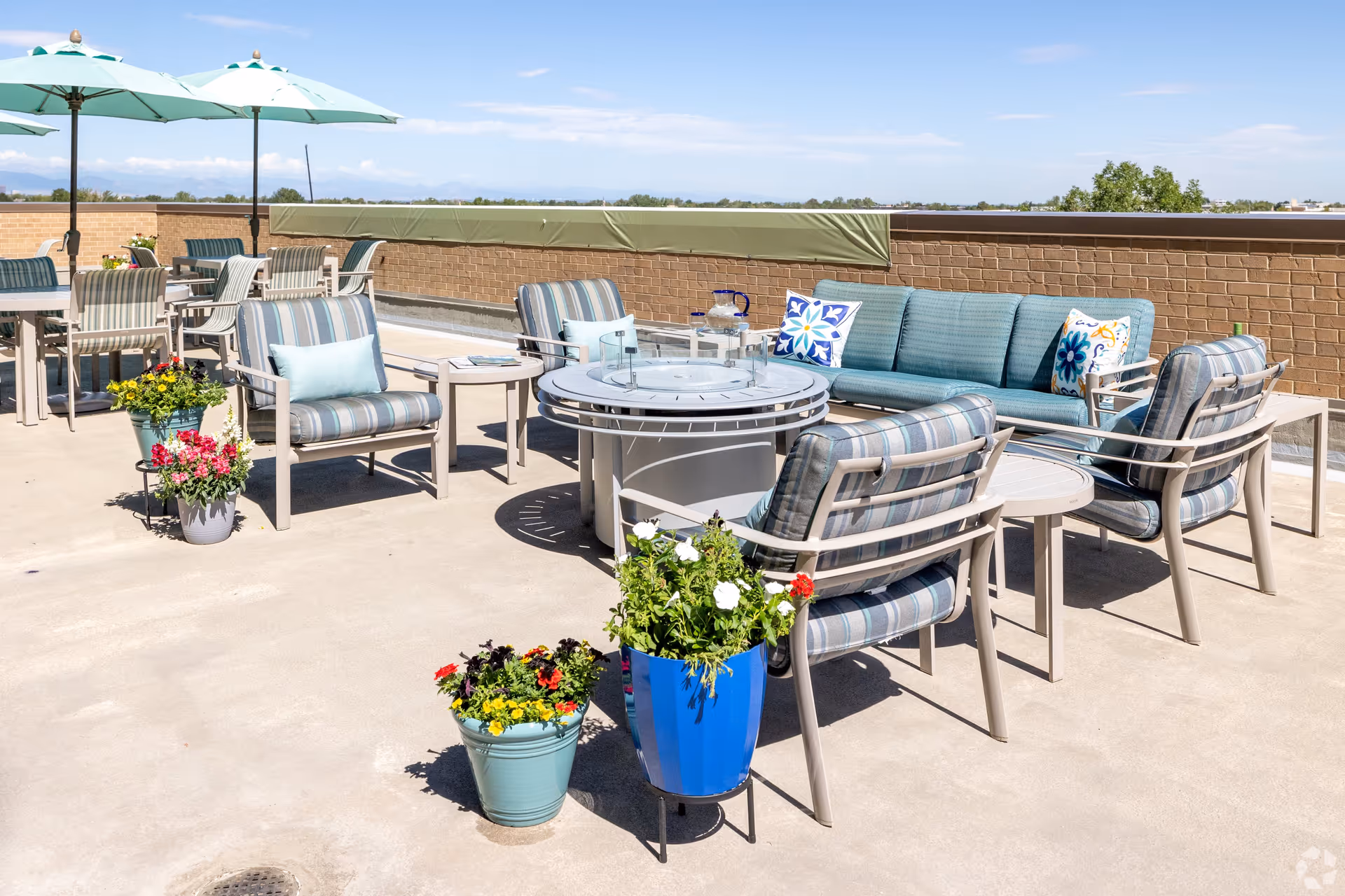 Outdoor patio area with cushioned chairs and a sofa arranged around a circular fire pit table. There are colorful flower pots placed near the seating area and tables with umbrellas in the background. The setting overlooks a low brick wall with a clear blue sky and distant mountains visible.