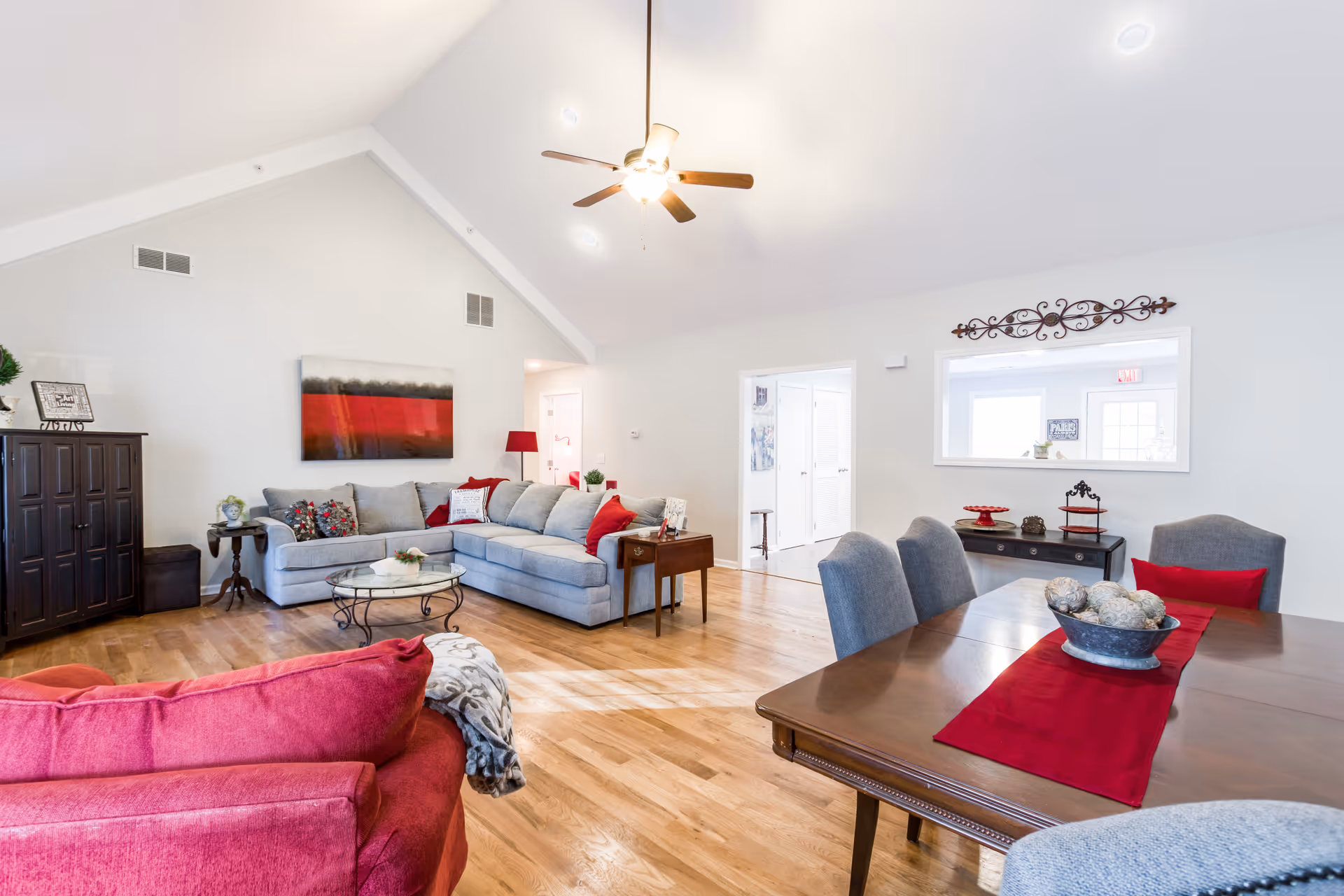 Bright and spacious living room with vaulted ceilings, featuring a gray sectional sofa with red and patterned pillows, a red armchair, a wooden dining table with gray chairs and a red runner, hardwood floors, a ceiling fan with lights, and decorative wall art and furniture.