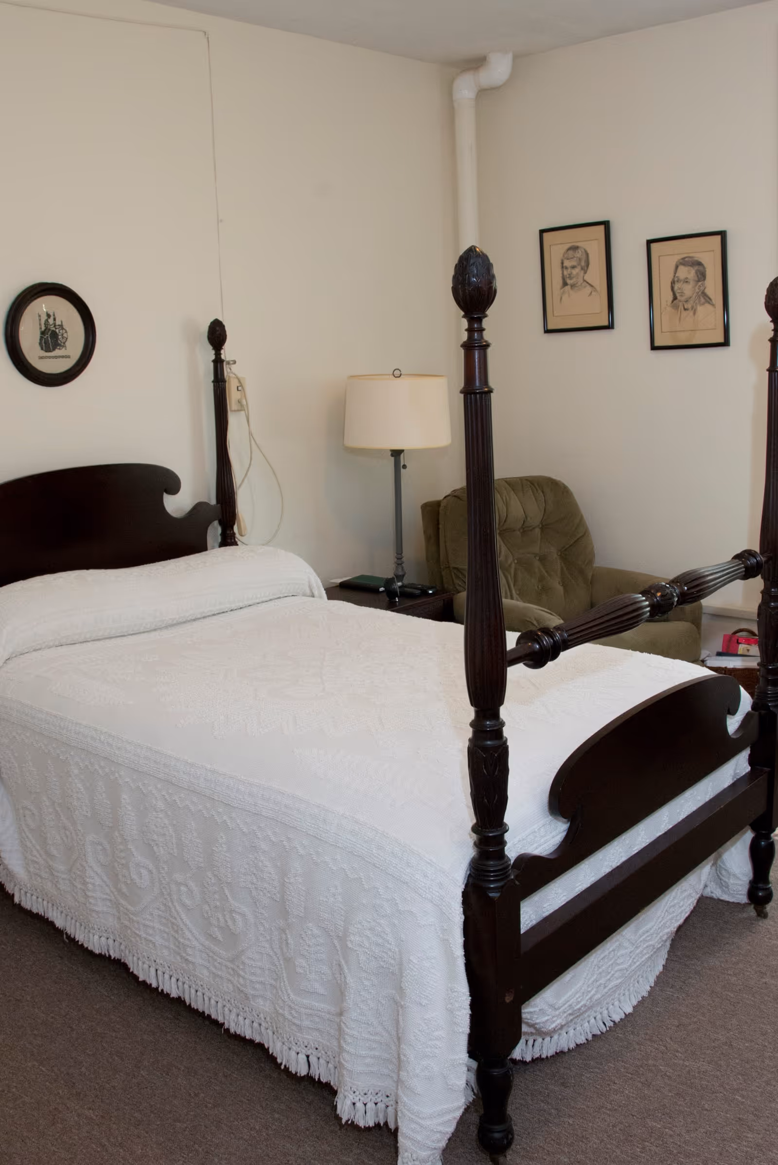 A bedroom with a dark wooden four-poster bed covered with a white textured bedspread. Next to the bed is a small table with a lamp and a green upholstered armchair. The walls are light-colored and decorated with three framed pictures.
