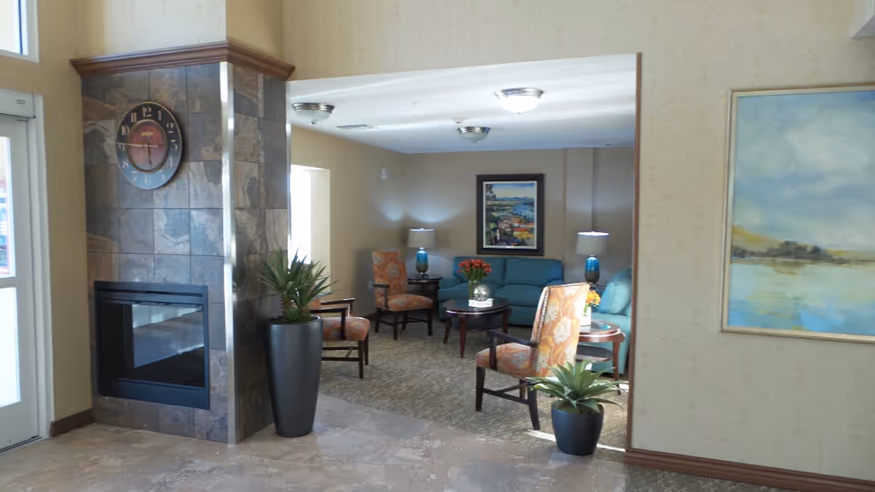Interior view of a senior living community lounge area with a tiled fireplace on the left, a clock above it, potted plants, and a seating area with a teal sofa, patterned armchairs, side tables with lamps, and framed artwork on the walls.