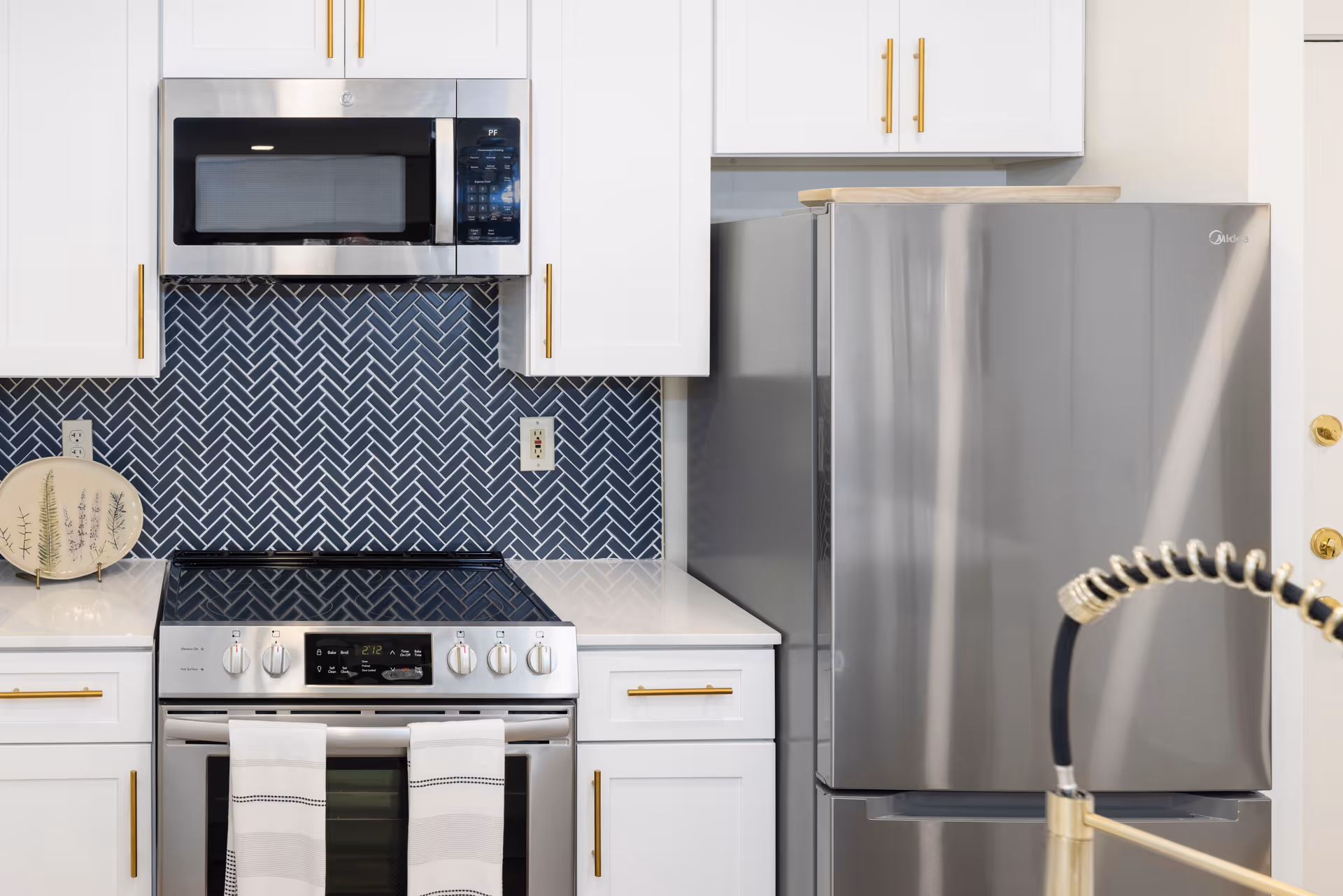 Modern kitchen with stainless steel refrigerator, stove and microwave, white cabinets and a dark herringbone tile backsplash.