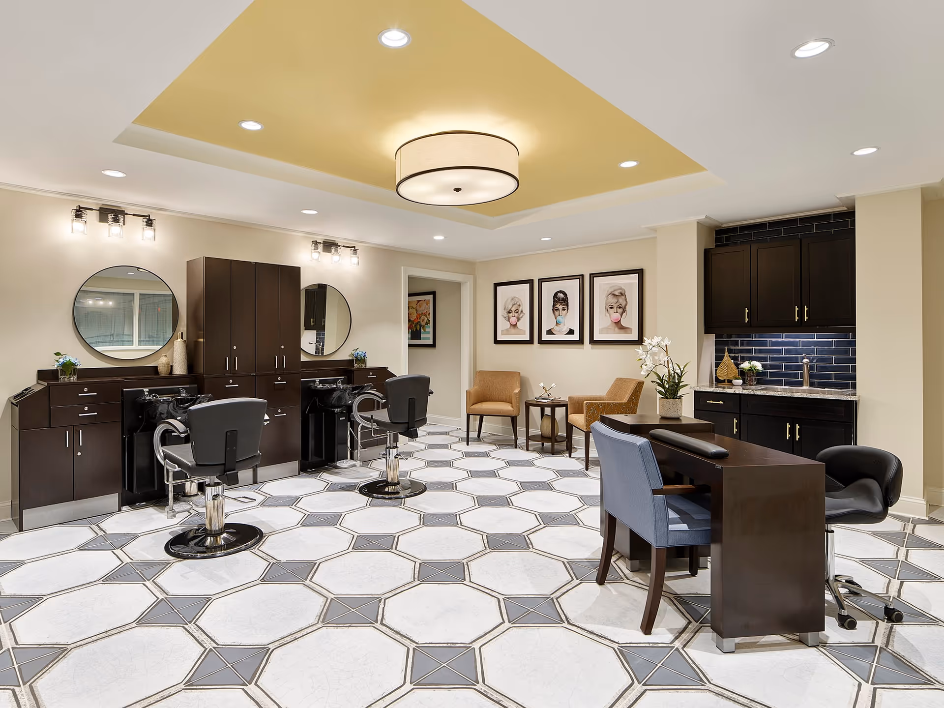 A modern salon area in a senior living facility featuring two black salon chairs in front of two round mirrors mounted on a wall with dark wood cabinetry. The floor has a geometric tile pattern in white and gray. To the right, there is a manicure table with two chairs, and in the background, two armchairs with a small table between them are placed under three framed artworks of women blowing bubble gum. The ceiling has recessed lighting and a central round light fixture with a yellow accent ceiling panel.