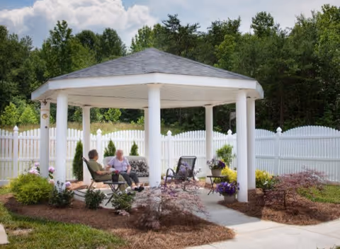 Two elderly people sitting and conversing under a white gazebo in a garden area surrounded by a white picket fence, with trees and shrubs in the background.