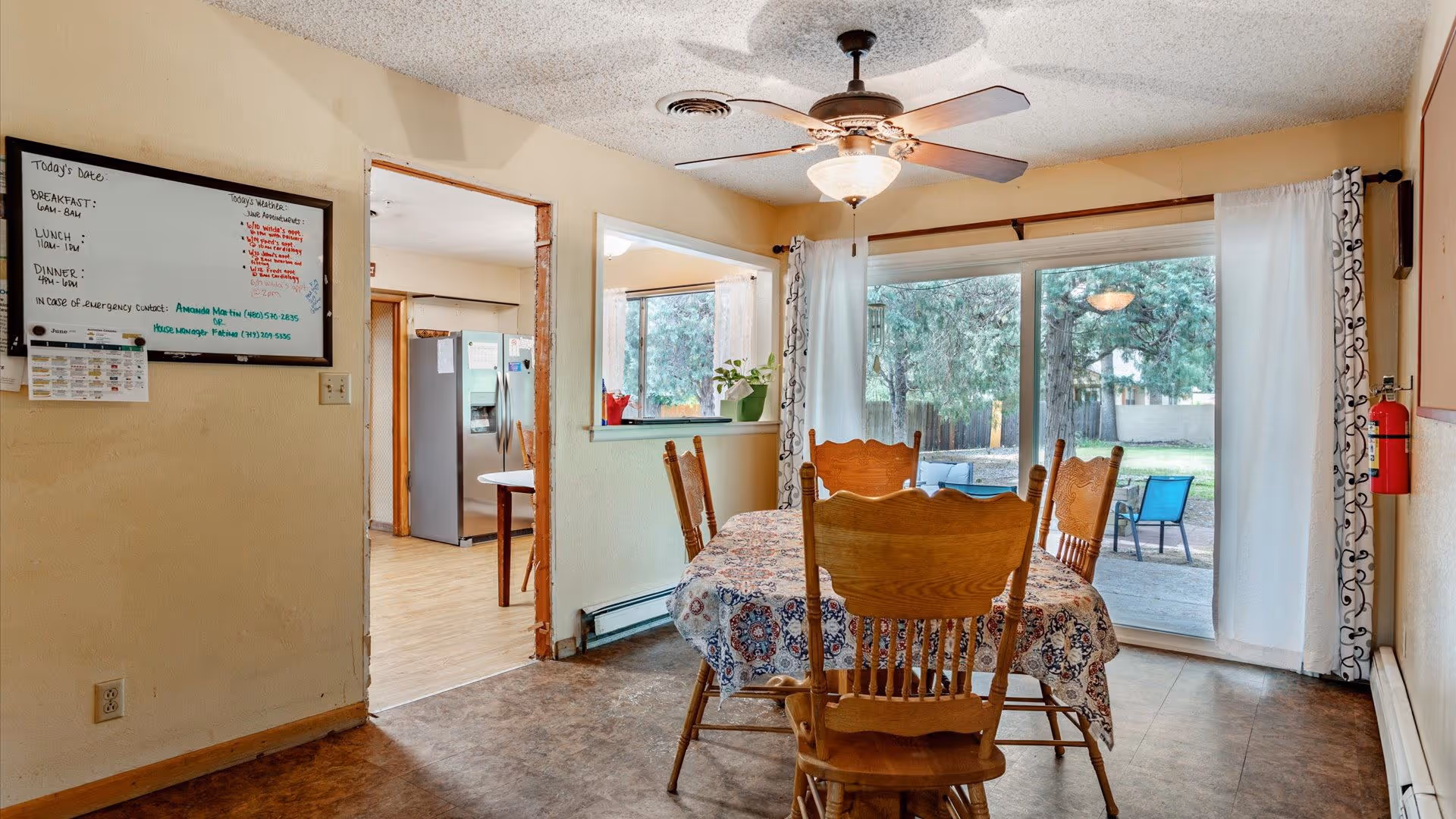 Dining area with a wooden table covered by a patterned tablecloth and four wooden chairs. A ceiling fan with a light fixture is above the table. Large sliding glass doors with white curtains open to a backyard with trees and outdoor seating. A whiteboard with daily meal information and emergency contact details is mounted on the wall to the left. The kitchen with a refrigerator is visible through an open doorway.