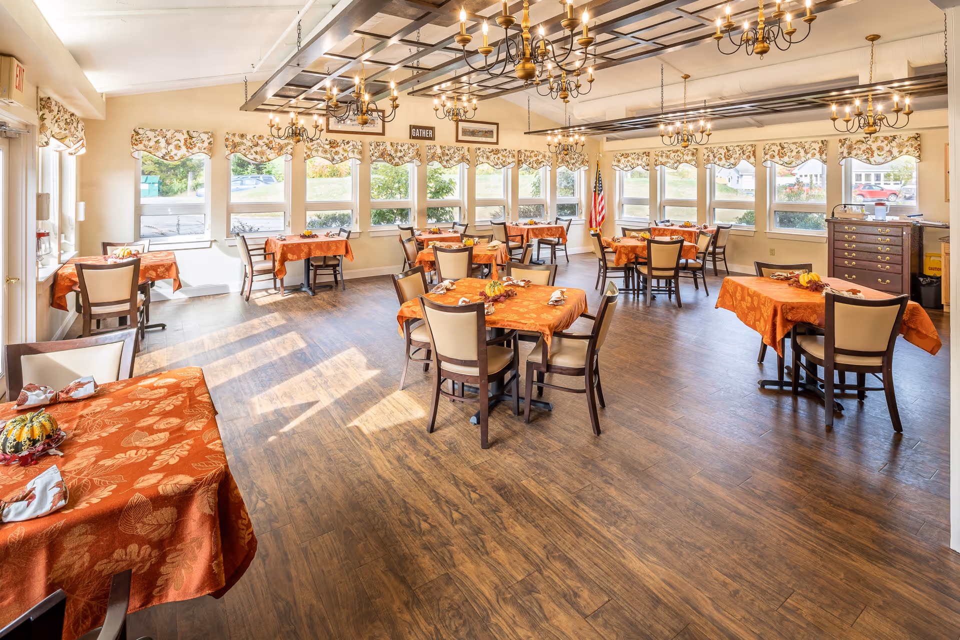 A bright dining room with multiple tables covered in orange tablecloths with autumn leaf patterns. Each table is set with napkins and small decorative pumpkins. The room has large windows with floral valances letting in natural light, wooden flooring, and several brass chandeliers hanging from the ceiling. An American flag stands in the corner near a wooden chest of drawers.