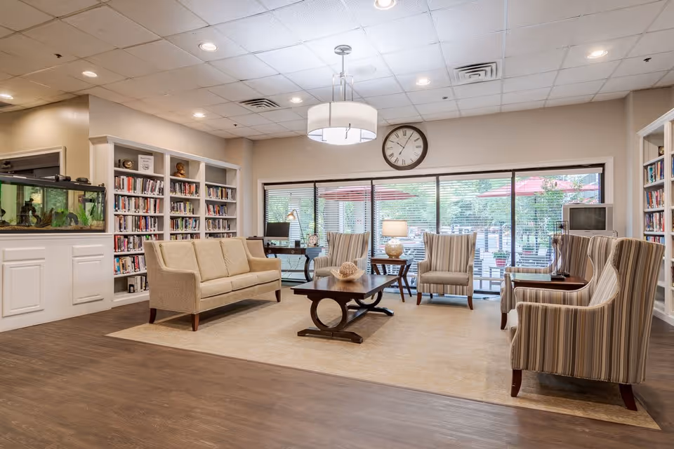 A bright and spacious living room area in a retirement community featuring a beige sofa, four striped armchairs arranged around a wooden coffee table, bookshelves filled with books, a large fish tank, a clock on the wall, and large windows with blinds overlooking an outdoor patio with red umbrellas.