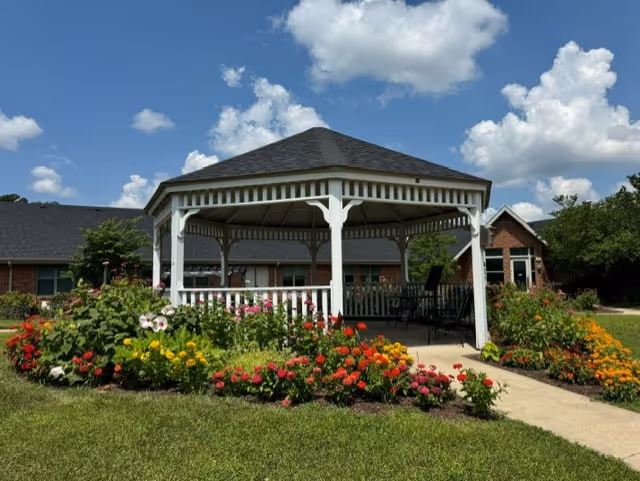 A white wooden gazebo surrounded by colorful flowerbeds on a lawn with a brick facility building and blue sky in the background.