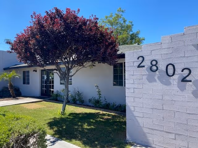Exterior view of a single-story building with white walls and a gray roof, featuring a tree with dark red leaves in front and a lawn with some shrubs. A white brick wall with the numbers 2802 is visible on the right side under a clear blue sky.