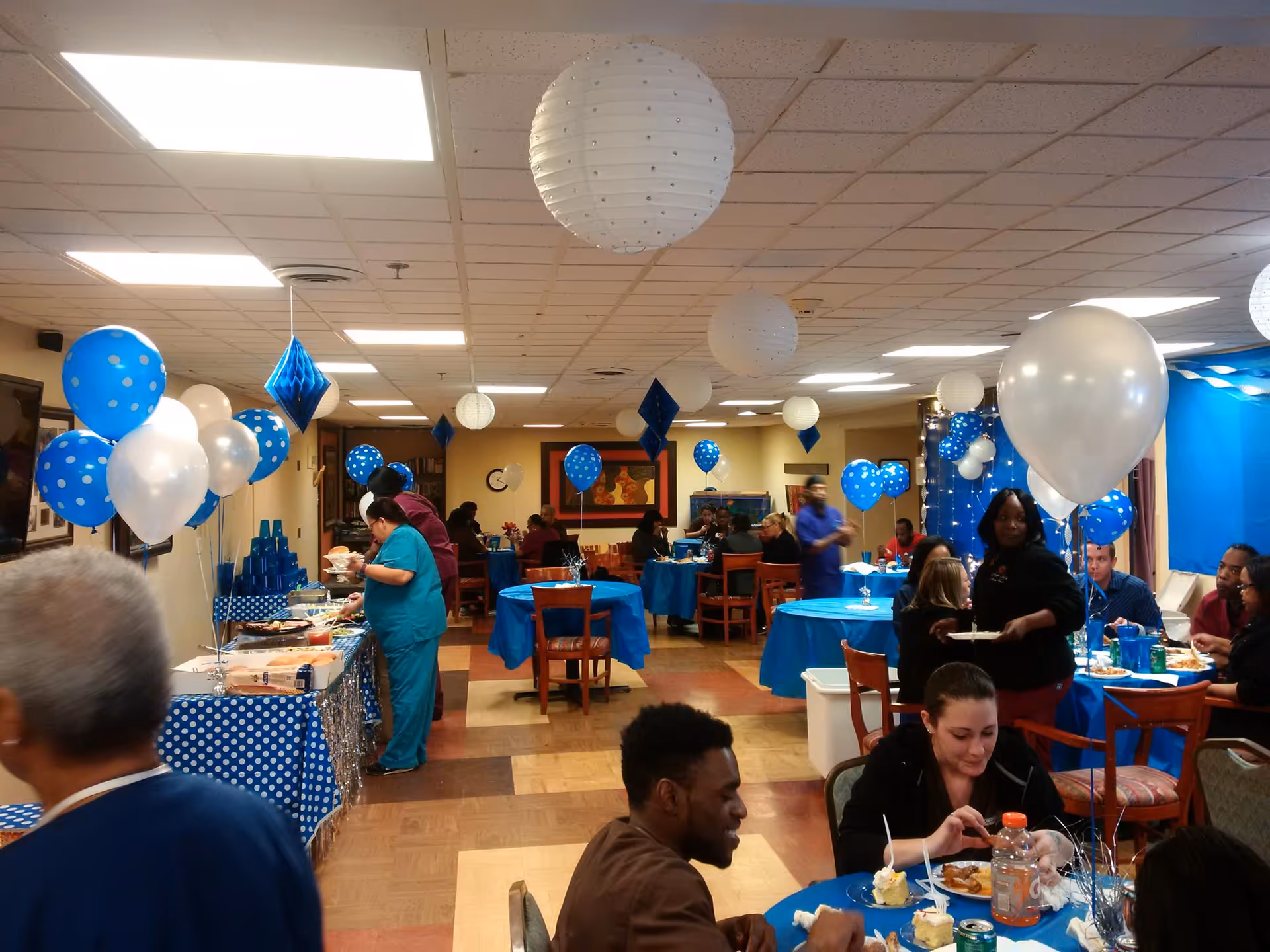 A decorated dining room with blue and white balloons and paper lanterns hanging from the ceiling. Several people are seated at tables covered with blue tablecloths, eating and socializing. A buffet table with food is set up on the left side, and staff members in scrubs are serving food. The room has a warm, festive atmosphere.