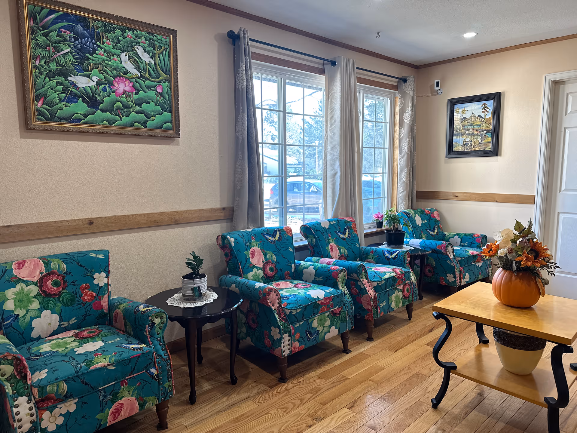 A cozy sitting area with four floral-patterned armchairs arranged along a wall with two large windows covered by light-colored curtains. There are two small black side tables with potted plants between the chairs, and a wooden coffee table with a pumpkin-shaped vase holding autumn flowers. The walls are decorated with two framed paintings, and the floor is wooden.